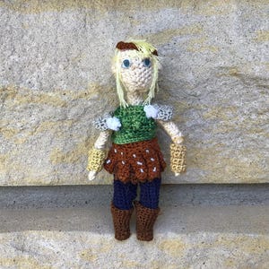 Puede incluir: Una muñeca tejida a ganchillo a mano con cabello de hilo rubio, ojos azules y una parte superior verde con detalles grises en los hombros. La muñeca lleva una falda marrón con costuras blancas, pantalones azul oscuro, puños amarillos y botas marrones.