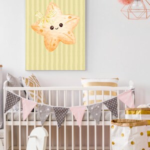 Könnte beinhalten: Ein Aquarelldruck eines lächelnden Seesterns mit gelber Schleife, vor einem hellgelb gestreiften Hintergrund. Die Kinderzimmerdekoration umfasst ein weißes Kinderbett mit einem Wimpelbanner in Rosa, Grau und Weiß.