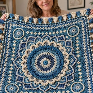 May include: A handmade, square crochet blanket in shades of blue and cream, featuring a central mandala design and decorative tassels along the edges. The blanket has a detailed, textured pattern and is held up by a person.