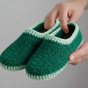 May include: A pair of green crochet slippers with a light green trim. The slippers are held by a person's hands. The slippers are a cozy, handmade item.