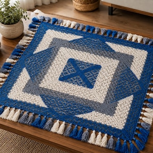 Può includere: Una coperta quadrata all'uncinetto con un motivo geometrico nei toni del blu reale, grigio ardesia e crema. Il design è composto da quadrati concentrici con un centro a forma di X, rifinito con una frangia decorativa di nappe alternate.