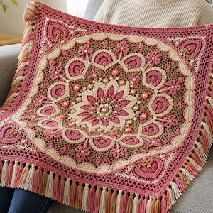 Peut inclure: Une couverture carrée au crochet présentant un motif mandala complexe dans des tons de vieux rose, crème et marron. Le design inclut des motifs floraux en relief, un travail ajouré complexe et est orné de nombreuses perles rondes. Les bords ont des franges.