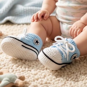 May include: A pair of baby crochet sneakers in light blue and white. The shoes have black accents, white laces, and a black star emblem on the side. The sneakers are designed to look like classic high-top sneakers.