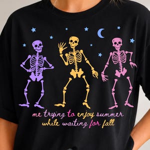 May include: A black short-sleeve t-shirt featuring a graphic of three dancing skeletons in purple, yellow, and pink. Above them are small blue stars and a crescent moon. The text reads: me trying to enjoy summer while waiting for fall.