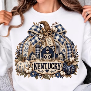 May include: A white sweatshirt featuring a large graphic of a pumpkin decorated with blue and white gingham patterns, a barn, and the text "KENTUCKY" and "home sweet home". The design includes cotton bolls, small blue flowers, and foliage.