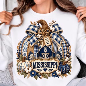 Könnte beinhalten: Ein weißes Sweatshirt mit Rundhalsausschnitt und einem großen Kürbis-Motiv, verziert mit blau-weißen Vichy-Karobändern, einer Scheune und Baumwollblüten. Der Schriftzug "MISSISSIPPI" ist in Blau auf einer cremefarbenen Plakette unten mittig.