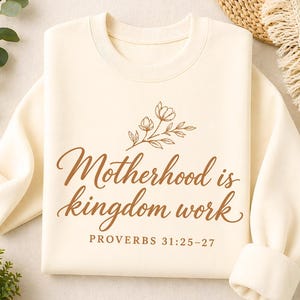 Può includere: Felpa color crema con la scritta "Motherhood is kingdom work" in marrone, accompagnata da un'illustrazione floreale. Il testo "PROVERBI 31:25-27" è presente. La felpa ha un girocollo e maniche lunghe.