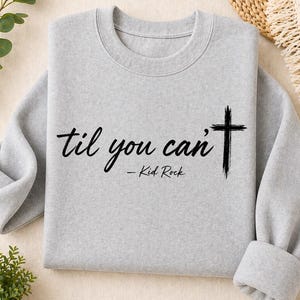 May include: A light gray sweatshirt with the text "til you can't" and a cross graphic. The text is in a handwritten style, with "- Kid Rock" below. The sweatshirt has a crew neck and long sleeves.