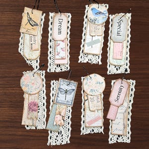 May include: A collection of handmade bookmarks with decorative paper tags, lace trim, and metal clips. The tags feature various designs, including butterflies, floral patterns, and the words "Dream," "Special," and "Serenity." The bookmarks are arranged on a dark wood surface.