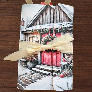 May include: A rectangular folded card featuring a winter scene of a wooden cabin with a bright red door, a festive wreath, and a stone path covered in snow. The card is tied with a light beige frayed ribbon and has small fabric tabs at the top.