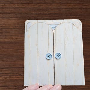 May include: A rectangular card featuring two arched, off-white doors with a vertical wood grain pattern. Each door is adorned with a small, light blue, four-hole button acting as a handle. The word "memories" is partially visible at the top center.