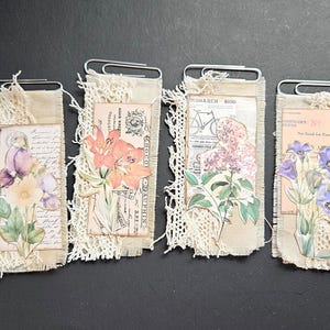 May include: Four handmade paper clip bookmarks. Each bookmark features a floral design with a vintage aesthetic. The designs include a bird, lilies, lilac, and blue flowers. The bookmarks are trimmed with lace and attached to paper clips.