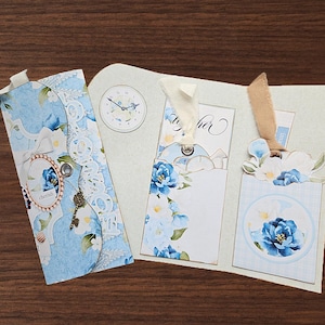 May include: A paper craft set featuring a blue floral patterned envelope with a small metal key charm and a button, alongside a folded card with two pockets containing floral tags, a clock illustration, and ribbons in cream and tan colors.