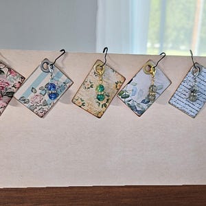 May include: Five diamond-shaped tags featuring diverse patterns including floral prints, blue stripes, and script text. Each tag is suspended by a black metal hook and includes a small dangling charm with a colored bead in red, blue, green, or grey.