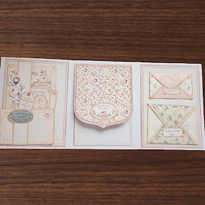 May include: A tri-fold paper card featuring vintage-style floral patterns and ephemera. The left panel displays layered tags and labels with French text. The center panel has a shield-shaped flap with floral print. The right panel shows two decorative envelopes.
