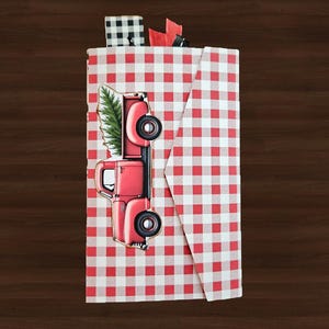 May include: A rectangular envelope featuring a red and white gingham pattern. A graphic of a red pickup truck carrying a green pine tree is centered on the front. The top edge shows small black and white checkered and red fabric ribbon accents.