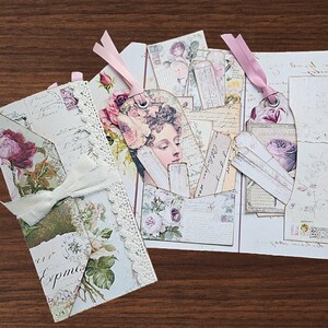 May include: A set of paper ephemera featuring floral and vintage script designs. The collection includes a folded card with white lace trim and a cream ribbon, alongside several tags with pink ribbons, all decorated with rose illustrations and text.
