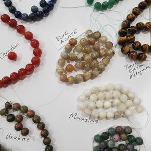 May include: Assortment of gemstone beads in various colors, including blue sodalite, red carnelian, green aventurine, brown tiger's eye, beige blue agate, white moonstone, green and brown unakite, and green and purple fancy jasper. Each bead strand is labeled.