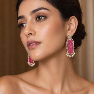 May include: Elegant pink gemstone earrings with a gold-colored filigree design. The earrings have a rectangular shape with a carved pattern and are adorned with small white pearls. The earrings are a statement piece.