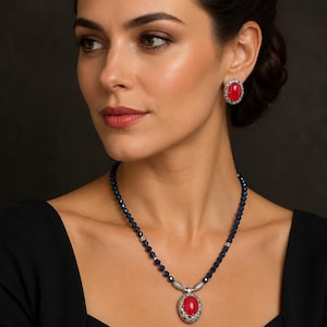 May include: A woman wearing a necklace and earrings set. The jewellery features a large oval red stone surrounded by a silver-coloured setting. The necklace has a dark blue beaded chain. The earrings match the necklace design.