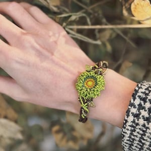 May include: A beaded bracelet featuring a large, green and yellow flower design. The flower is the focal point, with intricate beadwork creating a detailed floral pattern. The bracelet's band is brown and woven, with green leaf accents.