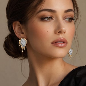 May include: Gold-colored earrings in the shape of melting clocks. The clock faces are white with black numbers and hands. The earrings have a surrealist design, with the clock faces appearing to drip down.