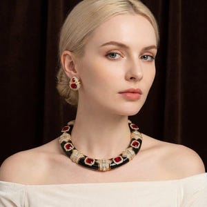 May include: A gold-toned necklace and matching earrings are worn by a woman. The necklace has black enamel segments with red gemstone accents and clear crystals. The earrings have a similar design. The woman is wearing a white off-the-shoulder top.
