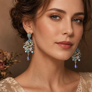 Puede incluir: Elegantes pendientes colgantes con diseño floral. Los pendientes presentan una combinación de cristales azules, turquesas y transparentes, con un cristal azul en forma de lágrima en la parte inferior. Los pendientes son dorados y son una pieza llamativa.