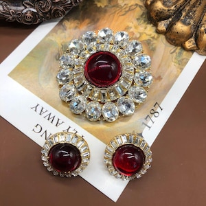 May include: A gold-toned jewelry set featuring a large brooch and matching earrings. The brooch and earrings have a central, round, deep red stone surrounded by clear, oval-cut crystals. The earrings and brooch are on a white paper with the text "NG AWAY 1787".