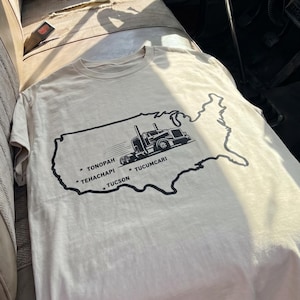 May include: A cream-colored short-sleeve t-shirt featuring a black graphic of the United States map with a semi-truck illustration. The design includes the text Tonopah, Tehachapi, Tucson, and Tucumcari with star markers placed across the map outline.