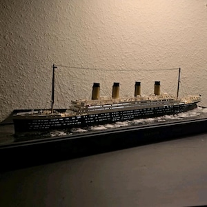 May include: A detailed scale model of the RMS Titanic ship featuring four yellow smokestacks, a black hull, and white upper decks. The model is illuminated with internal lights and is set on a base depicting waves with white foam detailing.