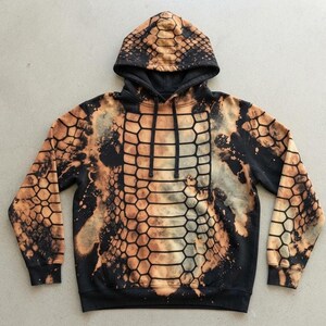 Op de afbeelding: Een zwarte hoodie met een gebleekt, bruinachtig slangenpatroon op de voorzijde, mouwen en capuchon. Het kledingstuk is voorzien van een kangoeroezak, lange mouwen met geribbelde boorden en zwarte trekkoorden bij de halslijn.
