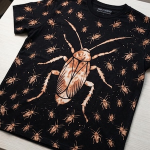 May include: A black short-sleeved t-shirt featuring a repeating pattern of small cockroach illustrations in a light brown, bleached effect. A large, central cockroach graphic dominates the front. The inner neck label reads VOID CLOTHING.