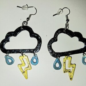 May include: A pair of dangle earrings featuring a black cloud-shaped frame with a textured surface. Each earring hangs with a central yellow lightning bolt charm and two light blue teardrop-shaped charms, all suspended from silver-toned hooks.