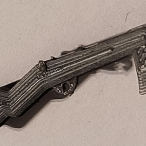 May include: A small, three-dimensional gray rifle figurine featuring a layered, ridged texture across its entire surface. The object displays a distinct stock, a central trigger area, and a vertical magazine component, all rendered in a monochromatic finish.