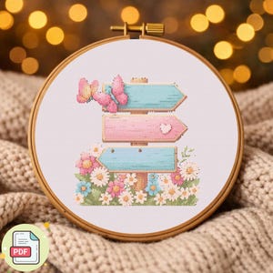 May include: A cross-stitch design in a wooden hoop features three directional signs in pastel blue and pink, adorned with butterflies and a heart. The signs are set against a backdrop of colorful flowers, creating a whimsical, nature-inspired scene.