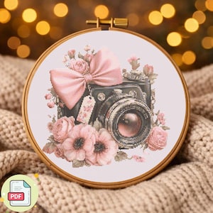 May include: Cross-stitch embroidery in a wooden hoop features a vintage camera design with pink flowers and a bow. The camera is black with a pink lens, and the flowers are various shades of pink. A small tag with floral details hangs from the camera.