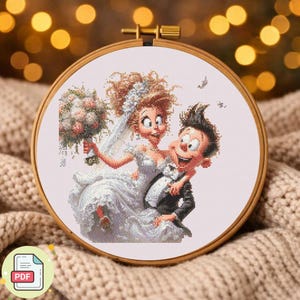 May include: A cross-stitch embroidery in a wooden hoop shows a cartoon bride and groom. The bride is in a white wedding dress and veil, holding a bouquet. The groom wears a tuxedo. The background is a soft, neutral colour.