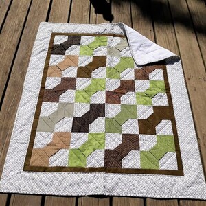 Op de afbeelding: Een groene, bruine en beige patchworkdeken met een witte achtergrond en een bruine rand. De deken is gemaakt van vierkanten en heeft een geometrisch patroon.