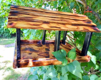 Puede incluir: Comedero para pájaros de madera con techo inclinado y base de bandeja. La madera tiene un acabado natural con detalles oscuros y carbonizados. El comedero está sostenido por postes de metal negro.