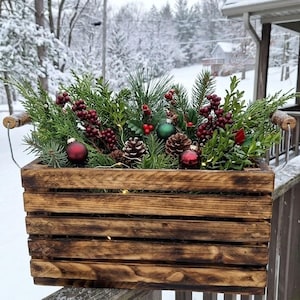 Peut inclure: Une caisse rectangulaire en bois teinté foncé avec des poignées en fil métallique, remplie de branches de conifères, de pommes de pin, de grappes de baies rouges et de petites boules de Noël rouges et vertes, agrémentée de guirlandes lumineuses.