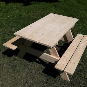 May include: A small, unfinished wooden picnic table with attached benches. The table has a rectangular top made of several planks and is supported by angled legs. The benches are also made of wooden planks. The table is set on a green grassy surface.