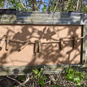 May include: A rustic wooden frame with a corkboard insert and a string of clothespins. The frame has a distressed green paint finish and is hanging on a tree branch.