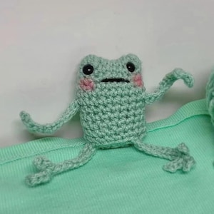 May include: A handmade crochet frog in light green yarn with black eyes and pink cheeks. The frog has outstretched arms and legs, and is sitting on a light green surface. The frog is a whimsical, handcrafted item.