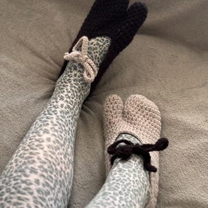 May include: A pair of split-toe crochet slippers, one in black and one in beige, each featuring a small decorative bow at the ankle. The slippers are worn over light-colored tights with a grey and white leopard print pattern throughout the fabric.