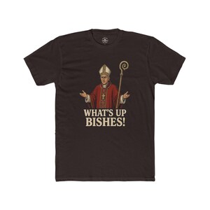 May include: Dark brown t-shirt featuring a graphic of a religious figure in red robes and a white hat, holding a staff. The text "WHAT'S UP BISHES!" is printed below the figure in a bold, vintage font.