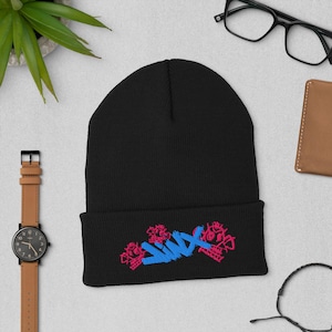 May include: A black beanie with a folded brim. The beanie features an embroidered design with pink skulls and blue text. Other items in the image include a watch, glasses, and a wallet.