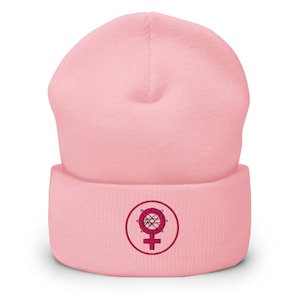 May include: A pale pink beanie hat with a turned-up brim. The hat has a circular embroidered emblem in a darker pink, featuring a female symbol. The beanie is made of a soft, knitted fabric.