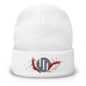 May include: A white beanie with a folded brim. The beanie features an embroidered design of a gray circle with three vertical lines and red splatter accents. The hat is made of a soft, knitted material.