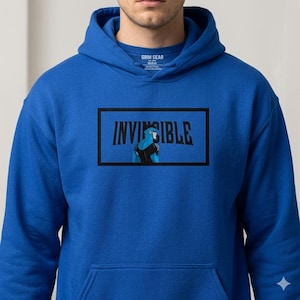 Könnte beinhalten: Königsblauer Kapuzenpullover mit einem Front-Grafik-Aufdruck „INVINCIBLE“ in schwarzen Blockbuchstaben. Die Grafik zeigt eine blau-schwarze Superheldenfigur in einem schwarzen Rechteck. Der Hoodie hat eine Kapuze mit Kordelzug und eine Vordertasche.